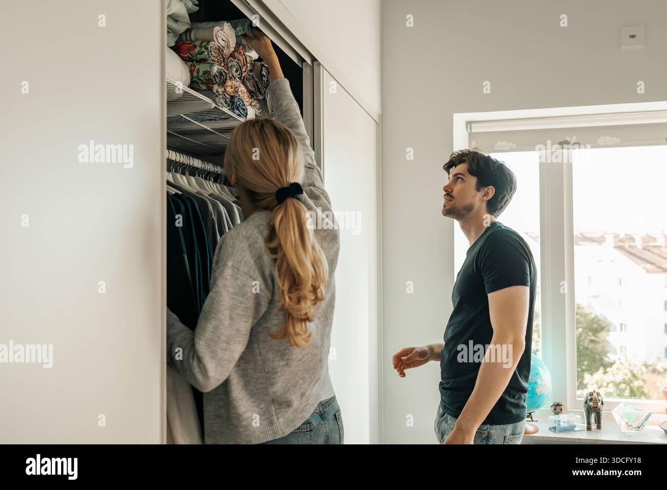 Junger Mann, der neben der Freundin steht und Bettlaken im Schrank in der neuen Wohnung organisiert Stockfoto