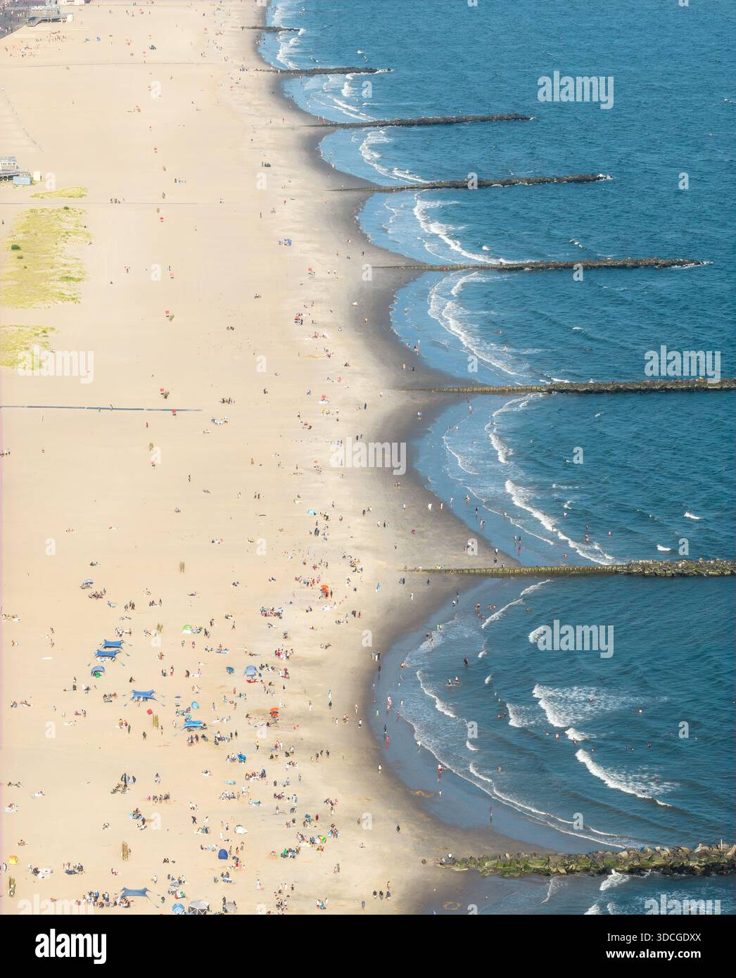 Blick aus der Vogelperspektive auf den Strand von Coney Island und die Wellenbrecher treffen sich in einem rhythmischen Tanz aus Sand und Meer, mit Menschenmassen am Ufer unter der Spätsommersonne. Stockfoto