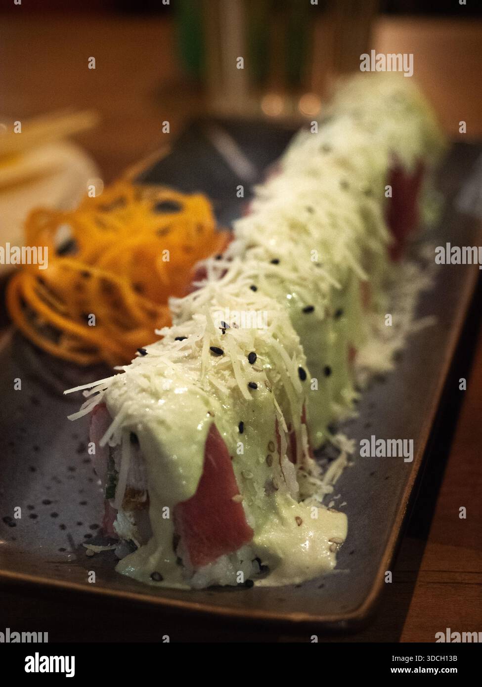 Ein Nahaufnahme-Foto zeigt eine lange Sushi-Rolle auf einer dunklen rechteckigen Platte. Das Brötchen wird großzügig mit einer hellgrünen, cremigen Sauce garniert Stockfoto