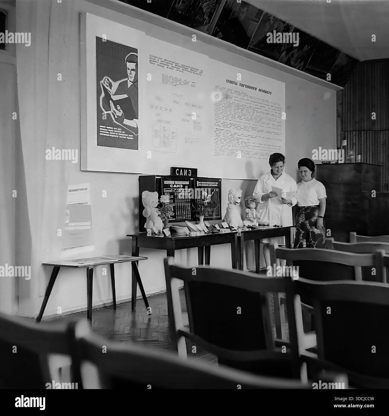 Slawjansk, Ukraine, UdSSR - 1980er Jahre: Zwei Frauen, wahrscheinlich Fabrikangestellte oder Parteiaktivisten, lesen ein Dokument in der Nähe einer Ausstellung von Konsumgütern (TNP) im Konferenzsaal des Armature-Isolator Plant (SAIZ). Eine Frau trägt einen weißen Labormantel. Hinter ihnen schmücken große Propagandaplakate mit dem Titel "Ratschläge für den Parteiaktivisten" die Wand. Die Szene umfasst hölzerne Stühle und eine Ausstellung von Spielzeug und Keramik, die das soziale und industrielle Leben im friedlichen Donbass widerspiegeln. Stockfoto