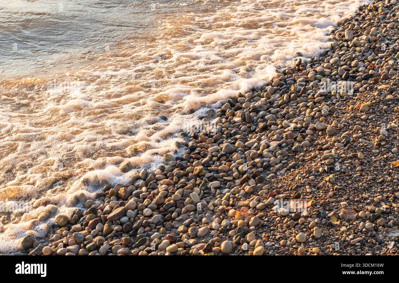 Sanfte Wellen sprudeln über glatte Kieselsteine entlang der Küste von Gimli, Manitoba, Kanada am Lake Winnipeg, die an einem Herbsttag in warmem natürlichem Licht leuchten Stockfoto