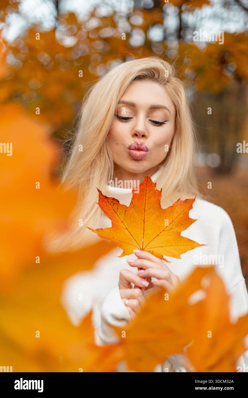 Frau küsst und hält ein großes Ahornblatt im goldenen Herbstwald. Ein positives trendiges Mädchen hält ein leuchtend bunt gelbes Ahornblatt. Herbststimmung und vib Stockfoto