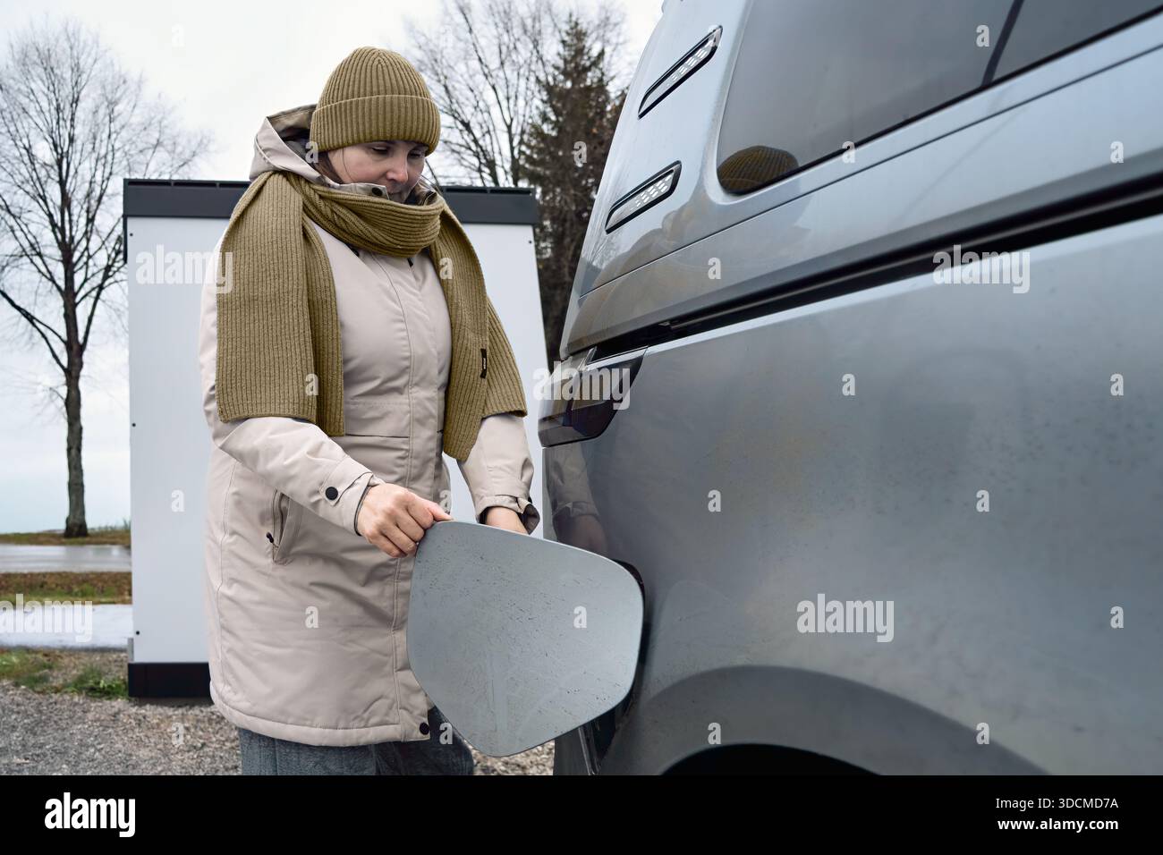 Leise Verantwortung prägt diesen Moment, als eine Frau mittleren Alters an einem kalten Tag den Ladeanschluss ihres Elektroautos im Freien öffnet und sich auf ewig vorbereitet Stockfoto