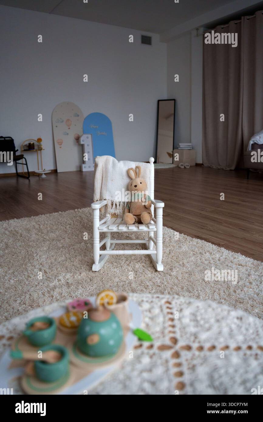 Gemütliches Kinderzimmer mit gefüttertem Häschen auf weißem Stuhl, Plüschteppich, Holzboden, dekorativen Requisiten und Spielzeug-Tee-Set für einen friedlichen Frieden Stockfoto