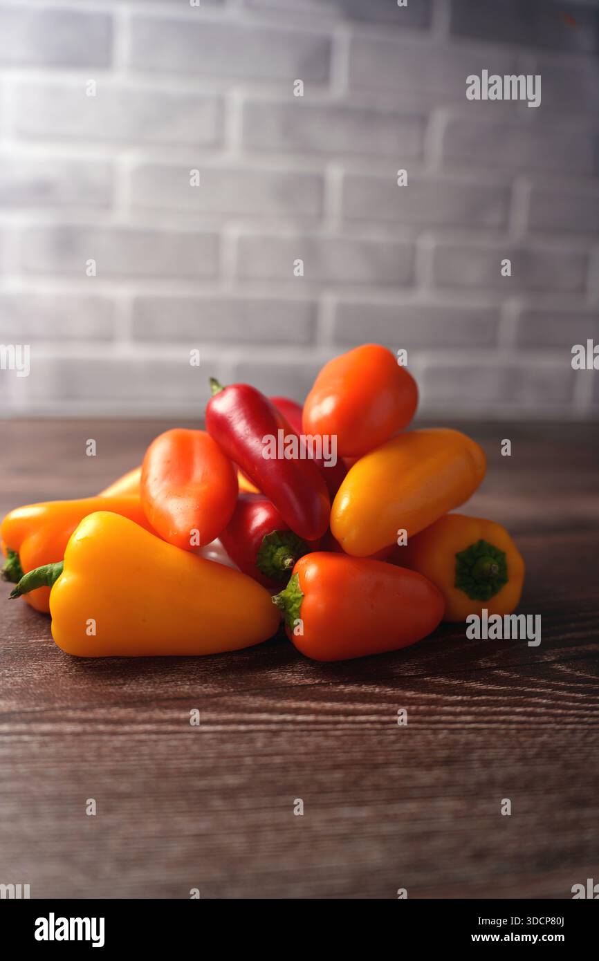 Ein lebhafter Haufen knallroter, oranger und gelber Mini-Paprika auf einer Holzoberfläche vor einem sanft beleuchteten Backsteinhintergrund, der eine frische und einladende Atmosphäre schafft Stockfoto