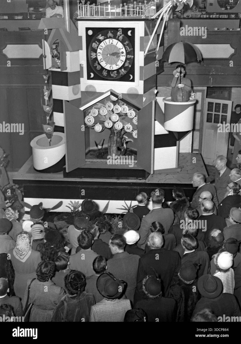 Eine Menschenmenge versammelte sich in Bingley Hall, Birmingham, West Midlands, England, Großbritannien, um 1953, um das Festival of Britain Guinness Clock zu sehen. Die beliebte Uhr zeigte „Guinness Time“, als animierte, von John Gilroy entworfene Tiere wie Tukane und ein Tierpfleger jede Viertelstunde – hier um 15:15 Uhr – auftraten. Die Uhr war ursprünglich bei den Feierlichkeiten 1951 im Battersea Park in London vertreten. Acht große und eine Miniatur-Version der mechanischen Uhr tourten mehrere Jahre lang durch britische Badeorte und Veranstaltungen – ein Vintage-Foto aus den 1950er Jahren. Stockfoto