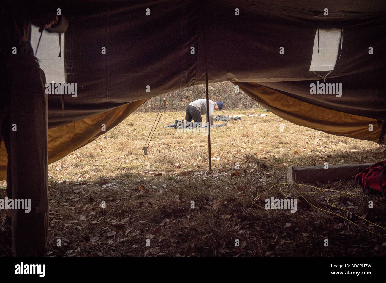 Blick aus dem Inneren eines Zeltes mit Blick auf eine Person, die ein Camp in einer Waldlichtung aufbaut, Überleben im Freien und Camping-Atmosphäre Stockfoto