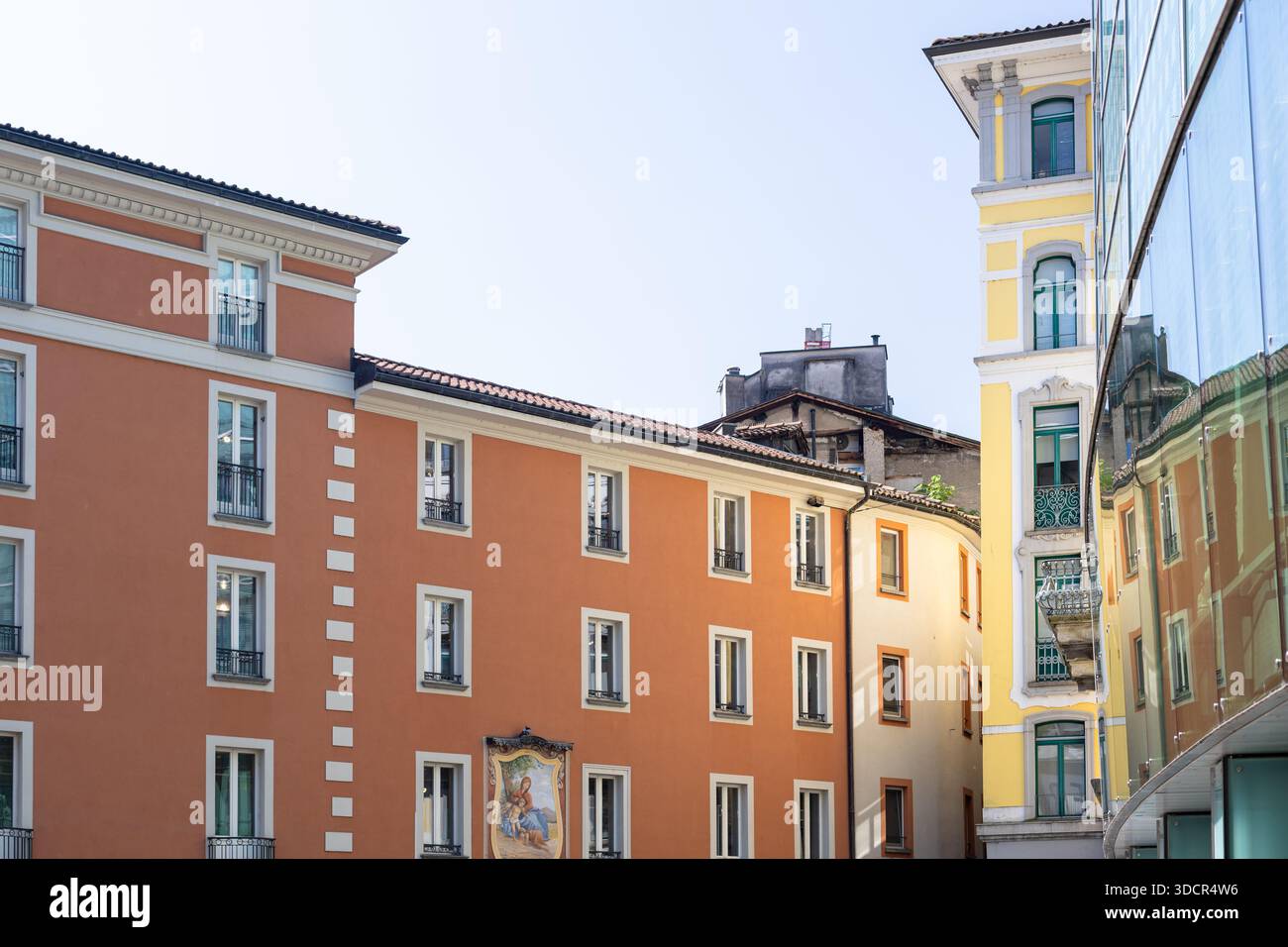 Sonnige europäische Straßenszene mit einem warmen orangen Apartmentblock, einem Gebäude mit gelben Akzenten und einer eleganten Glasfassade, die die umliegenden Archi reflektiert Stockfoto