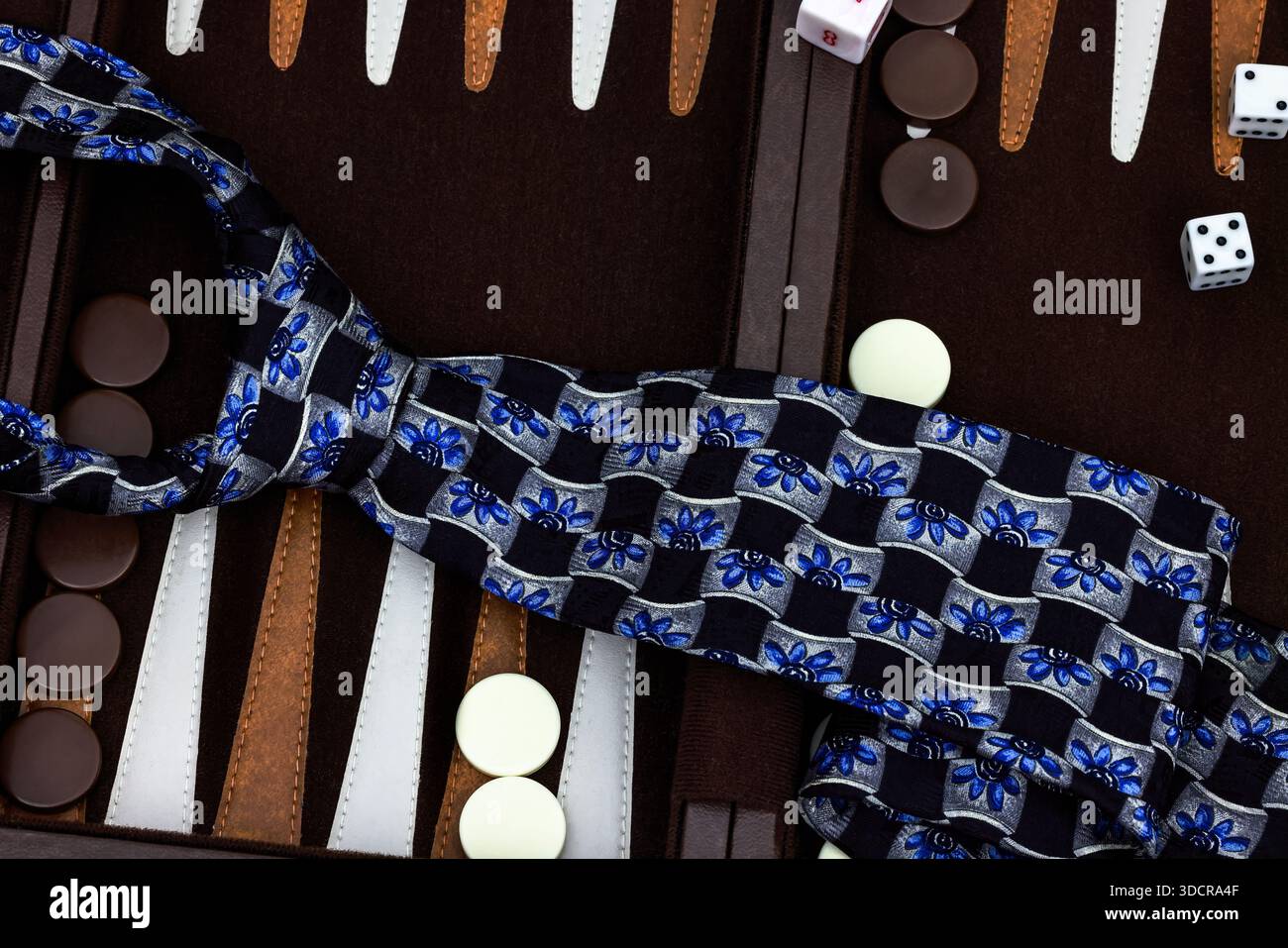 Nahaufnahme einer bunten Krawatte aus Seide in Schwarz, Blau und Silber, die über Backgammon-Stücke und Würfel hängt und elegante Mode mit Freizeit verbindet Stockfoto