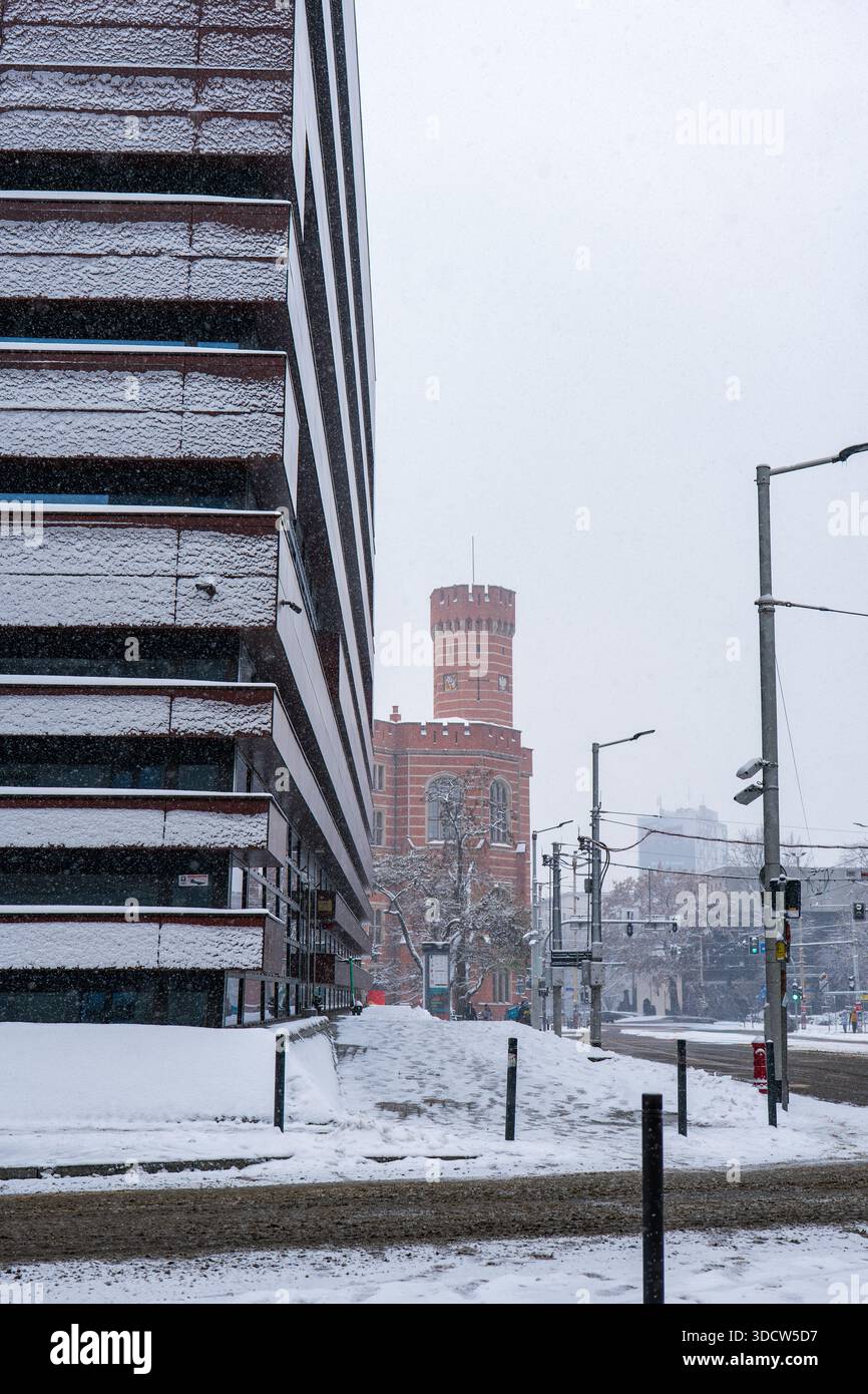 Frischer Schnee liegt auf einem modernen Büro aus Glas und Metall in Breslau, hinter dem sich ein roter Backsteinturm im neogotischen Stil erhebt. Schnee fällt über die Straßenbahnleitungen, der Verkehr ist leicht Stockfoto