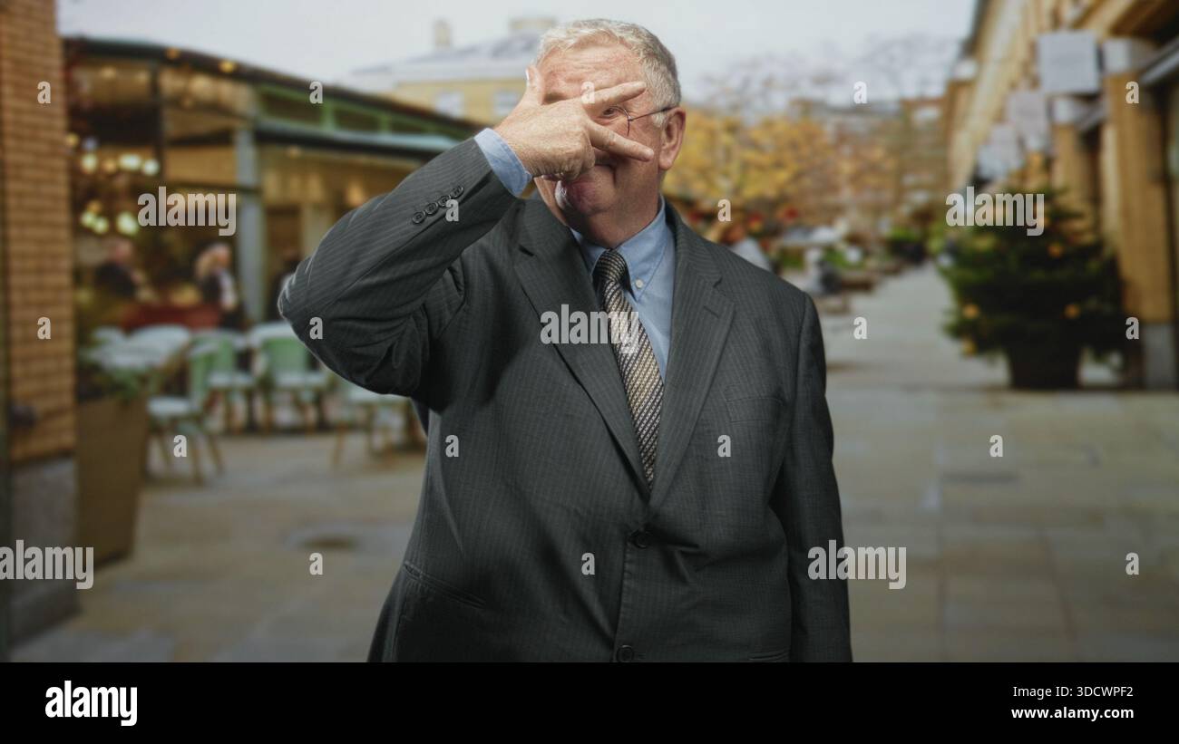 Mann im Anzug macht eine V-Form mit den Fingern über dem Auge auf einer Fußgängerzone, die von Geschäften und einem Weihnachtsbaum gesäumt ist; schelmisch. Stockfoto