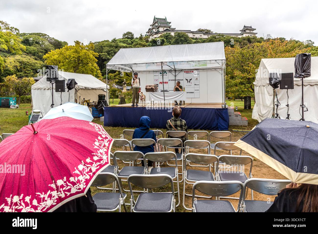 Ein Mann spricht mit einem ausgebildeten Spürhund auf einer Bühne vor einem Publikum auf dem Festplatz von Wakayama Castle, Japan, ein Banner mit der Aufschrift "PEPPY" hängt hinter ihm. Zuschauer beobachten die Vorführung unter Regenschirmen. Toyotomi Hideyoshi ließ die Burg 1585 errichten; der heutige Bergfried ist ein Wiederaufbau aus Stahlbeton von 1958. Stockfoto