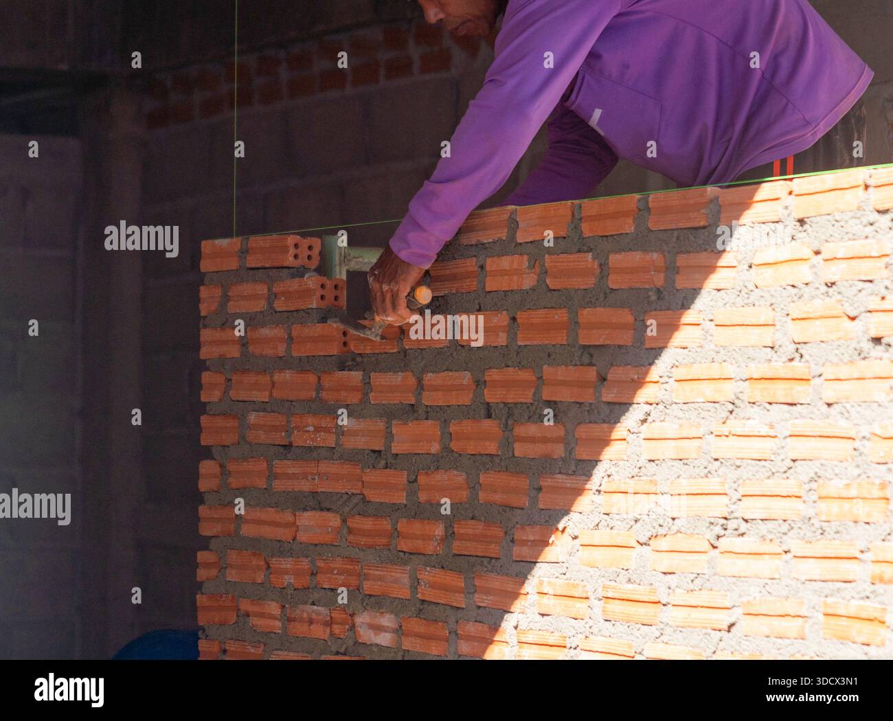 Ein maurer in einem lila Hemd verwendet eine Kelle, um Mörtel auf eine Ziegelwand aufzubringen. Dabei verwendet er eine grüne Führungslinie, um die Präzision während der Konstruktion zu verbessern. Stockfoto