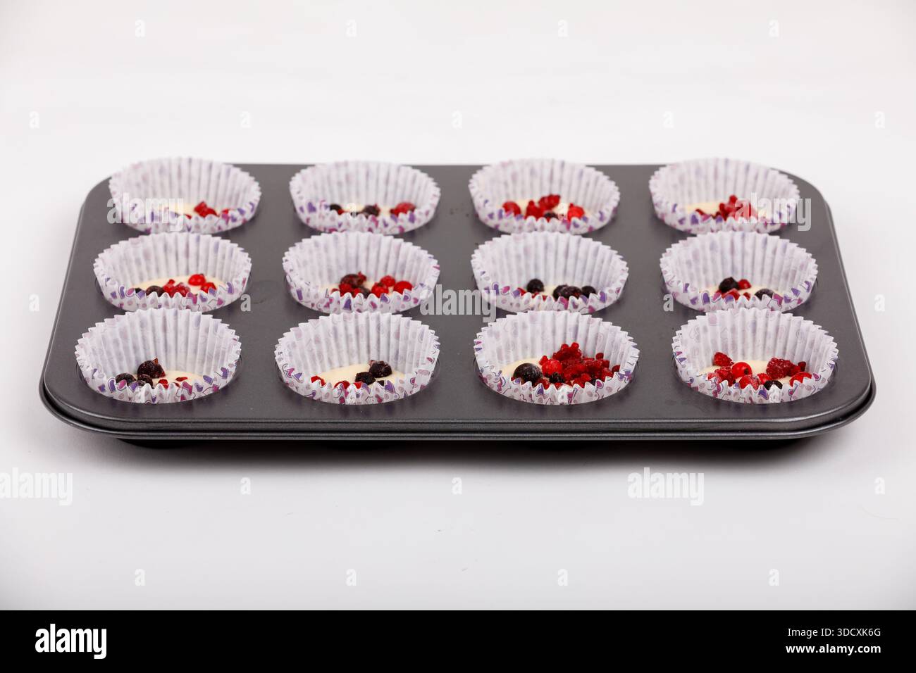Nahaufnahme eines Muffintabletts mit Cupcake-Einlagen gefüllt mit Teig und gemischten Beeren, die bereit sind, süße Leckereien zu Hause zu backen. Stockfoto
