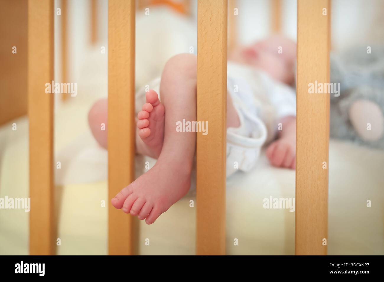 Ein Baby liegt in einem Kinderbett mit kleinen Füßen zwischen den Holzstäben. Die Einstellung ist hell und zeigt weiche Betten an. Das Baby ruht sich gut aus. Stockfoto