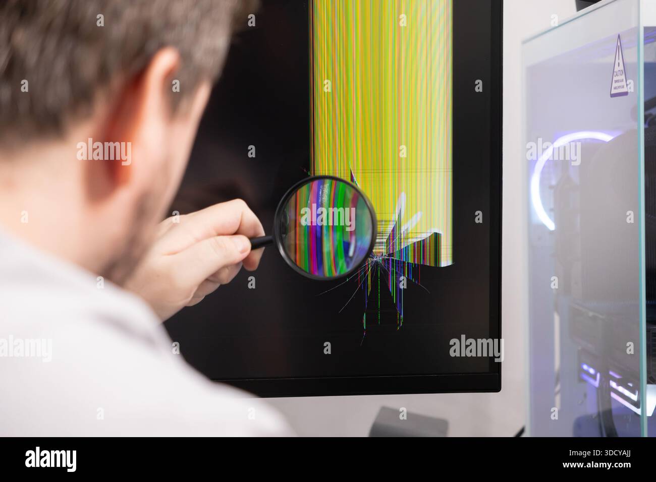 Nahaufnahme einer Person, die mit einer Lupe einen stark gerissenen Computermonitor mit leuchtenden toten Pixeln untersucht, die Elektronik hervorheben Stockfoto