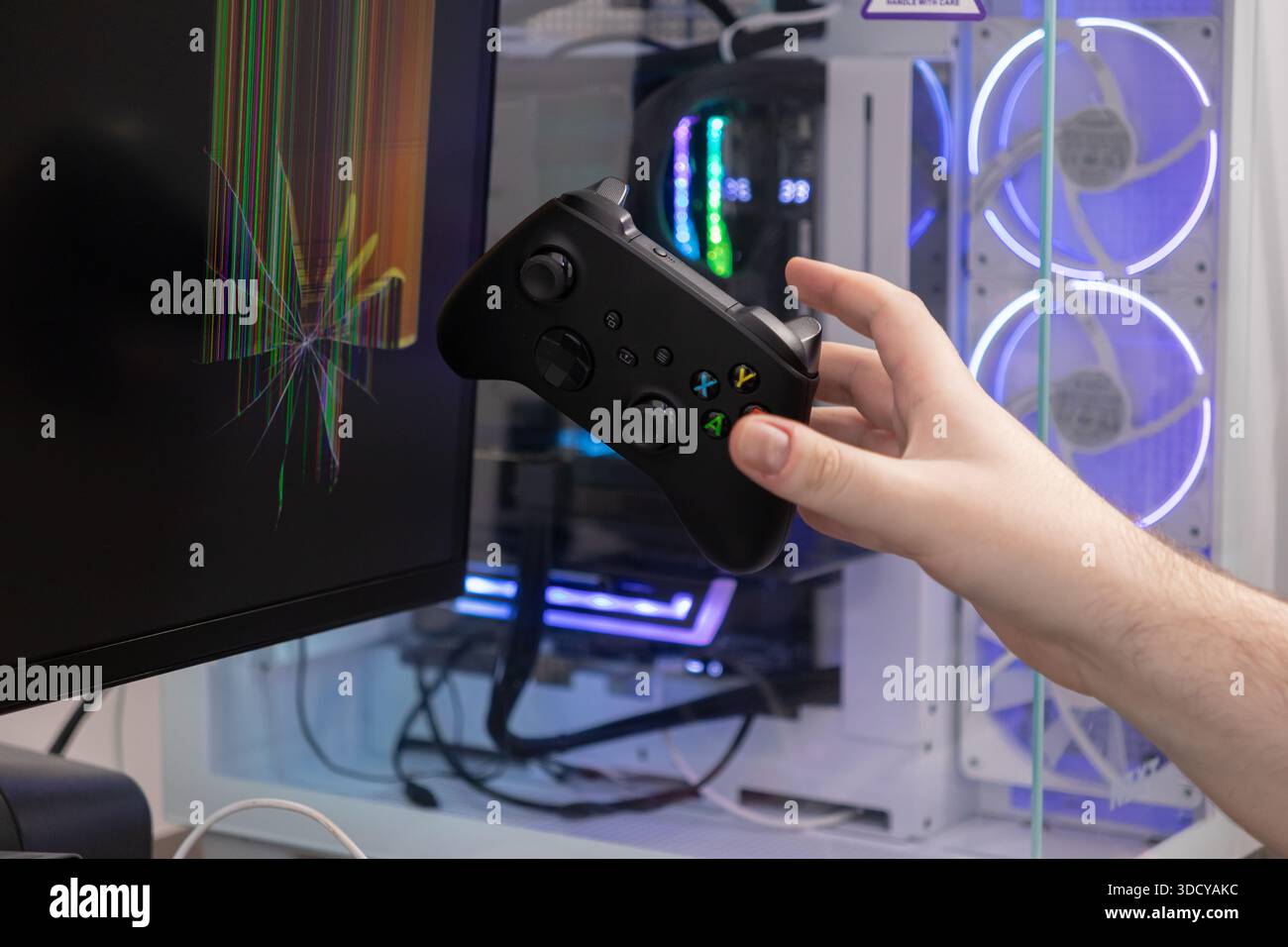 Die Hand greift einen Videospiel-Controller in der Nähe eines stark kaputten Computermonitors mit gebrochenen Pixeln, wodurch Schäden an der Gaming-Hardware und die Frustration der Spieler hervorgehoben werden Stockfoto