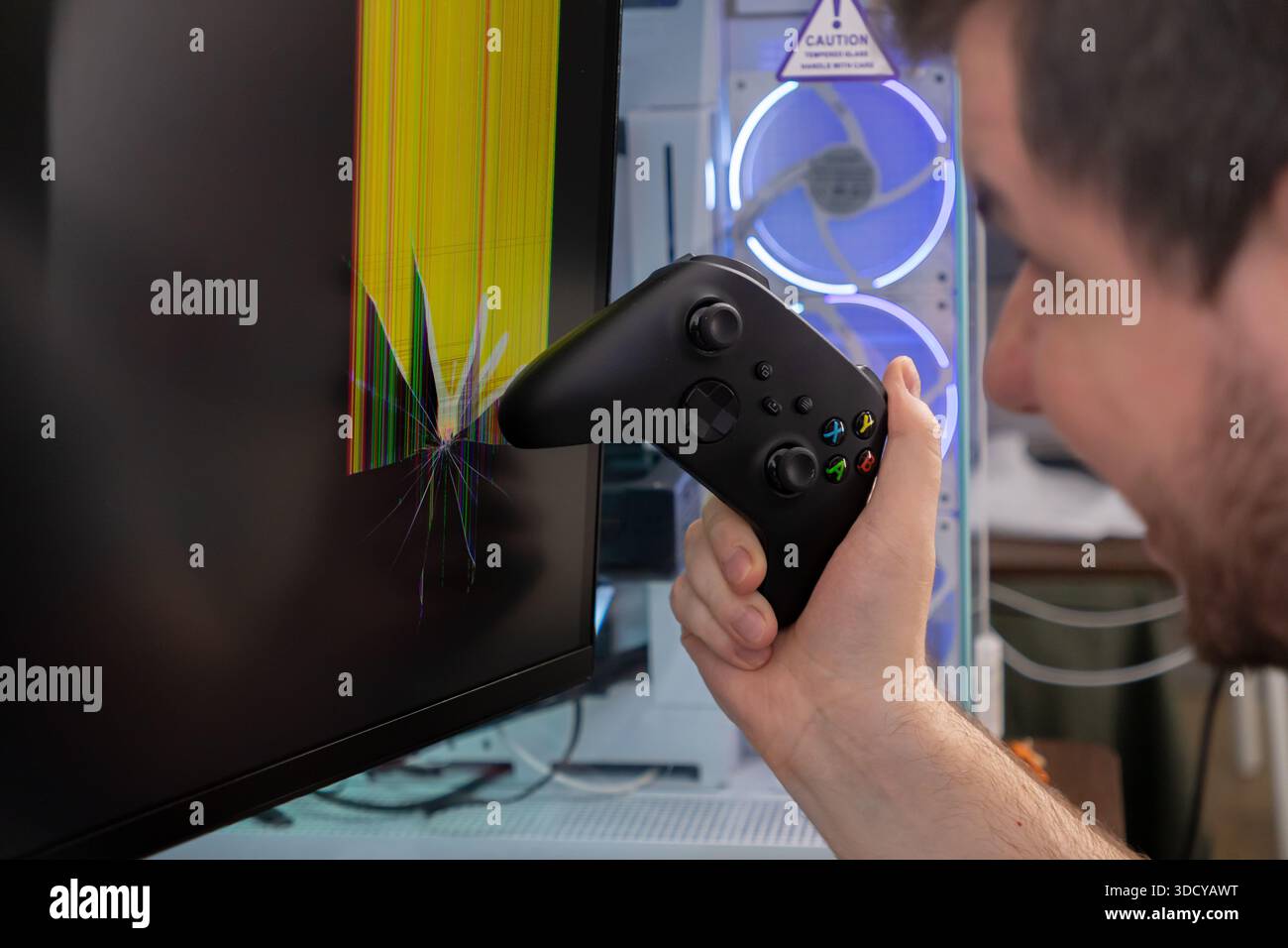 Mit der Hand wird das schwarze Gamepad in der Nähe eines zerschmetterten Computerbildschirms mit bunten, glitschigen Pixeln gefasst, die Spielwüte, technische Probleme und kostspielige Inhalte betonen Stockfoto