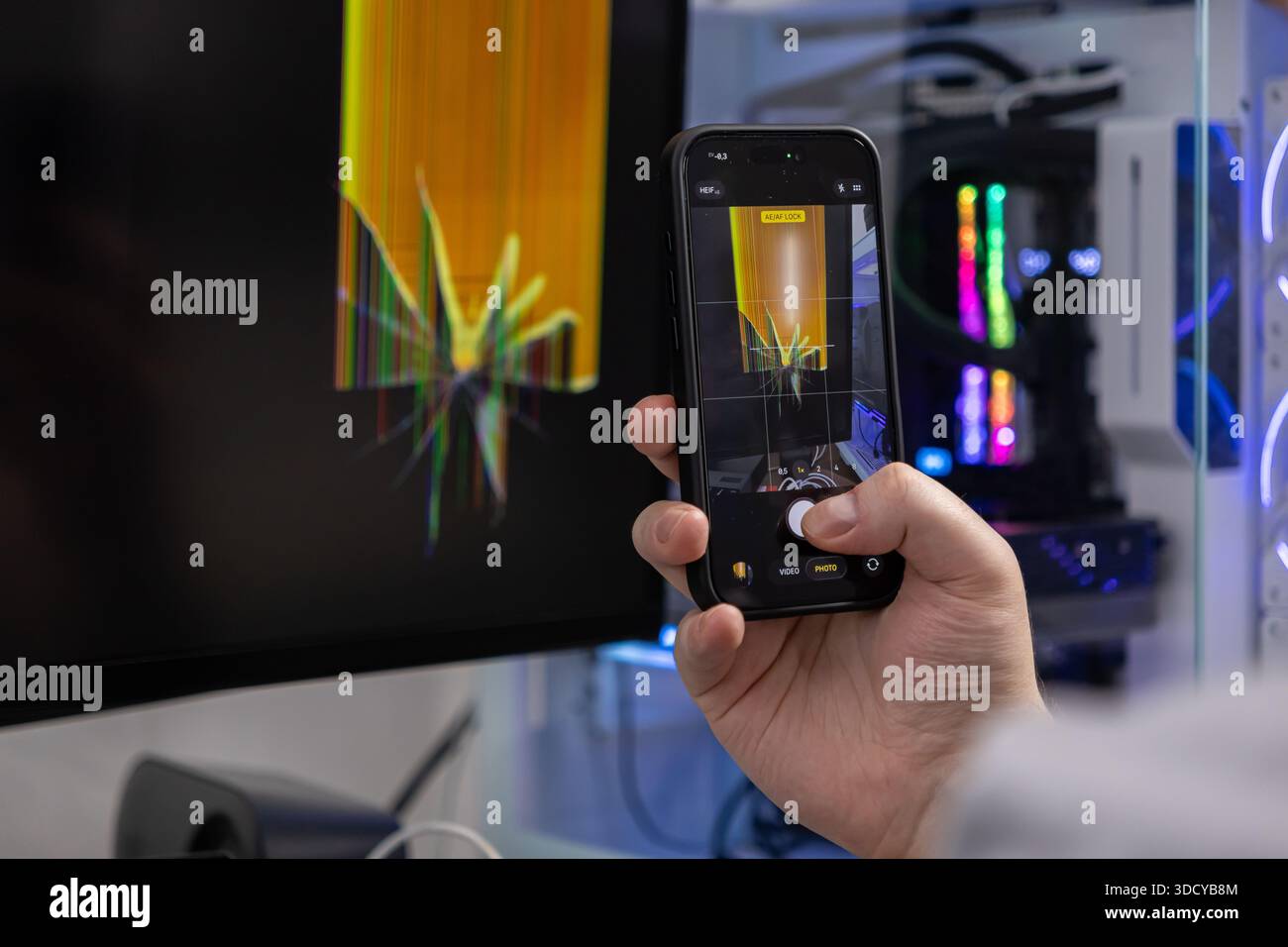 Nahaufnahme einer Hand, die ein Telefon hält, dokumentiert den schwer beschädigten Desktop-Computer-Monitor, der eine Explosion von bunten Pixeln zeigt, die für r wichtig sind Stockfoto