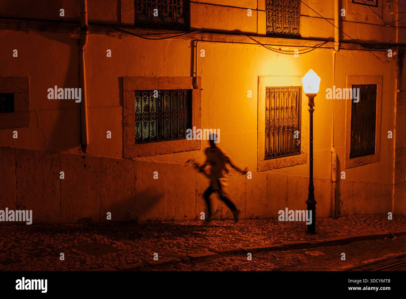 Person, die nachts an einer Straßenlaterne auf einer Kopfsteinpflasterstraße in Lissabon, Portugal, vorbeirennt. Stockfoto
