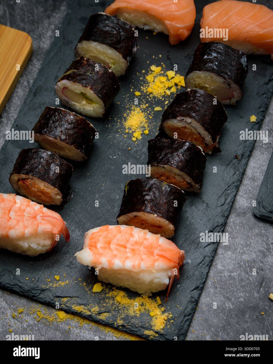 Nahaufnahme mit verschiedenen Sushi-Brötchen und Garnelen-Nigiri, die in ordentlichen Reihen auf einem dunklen Schieferbrett angeordnet sind, mit japanischem Gourmet-Hintergrund für Menüs. Stockfoto