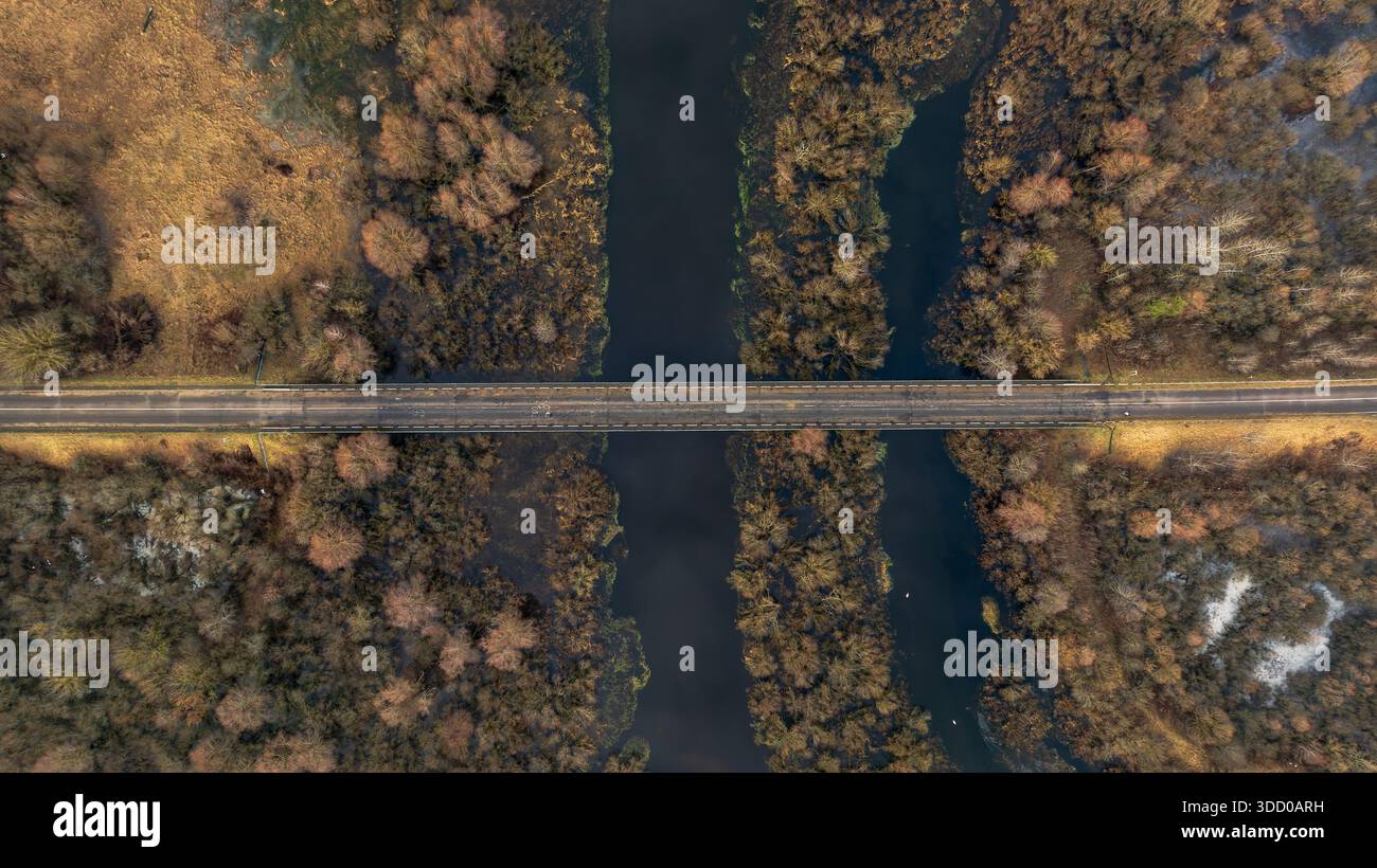Die Luftaufnahme zeigt eine gerade zweispurige Brücke über einen dunklen, langsamen Fluss in Belarus. Wintermoor und Blattlose Wälder umgeben dünnes Eis und Frost in weichen Farben Stockfoto