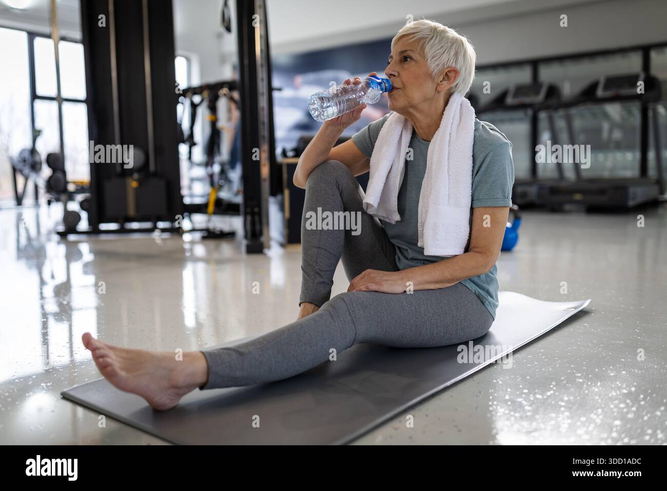 Eine Frau sitzt auf einer Matte im Fitnessstudio und trinkt nach dem Training Wasser. Sie hat ein Handtuch um den Hals und sieht entspannt aus. Stockfoto
