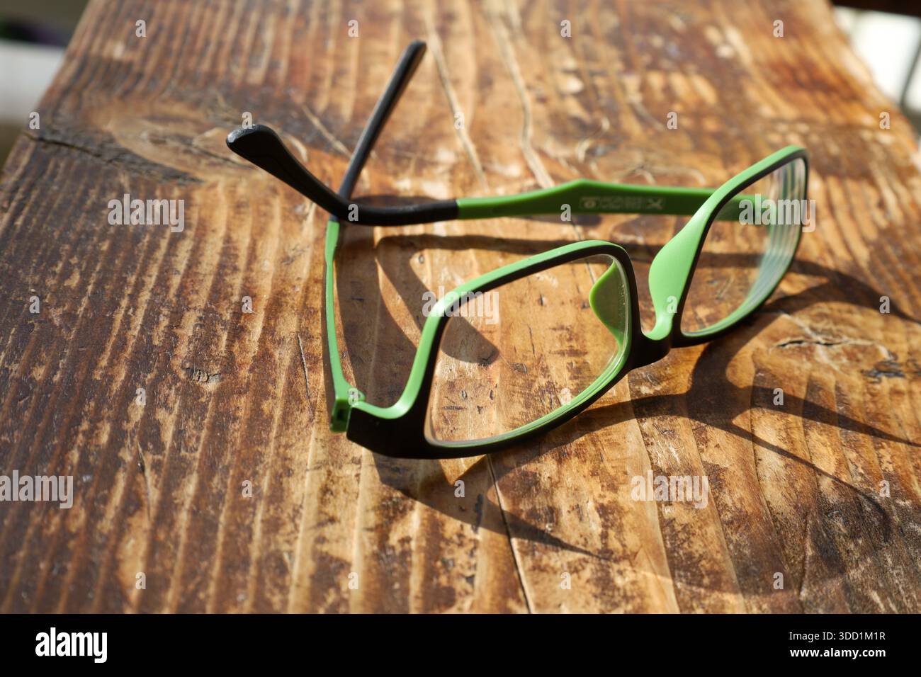 Eine modische grüne und schwarze Brille liegt auf einem rustikalen Holztisch, beleuchtet von der warmen Nachmittagssonne. Stockfoto