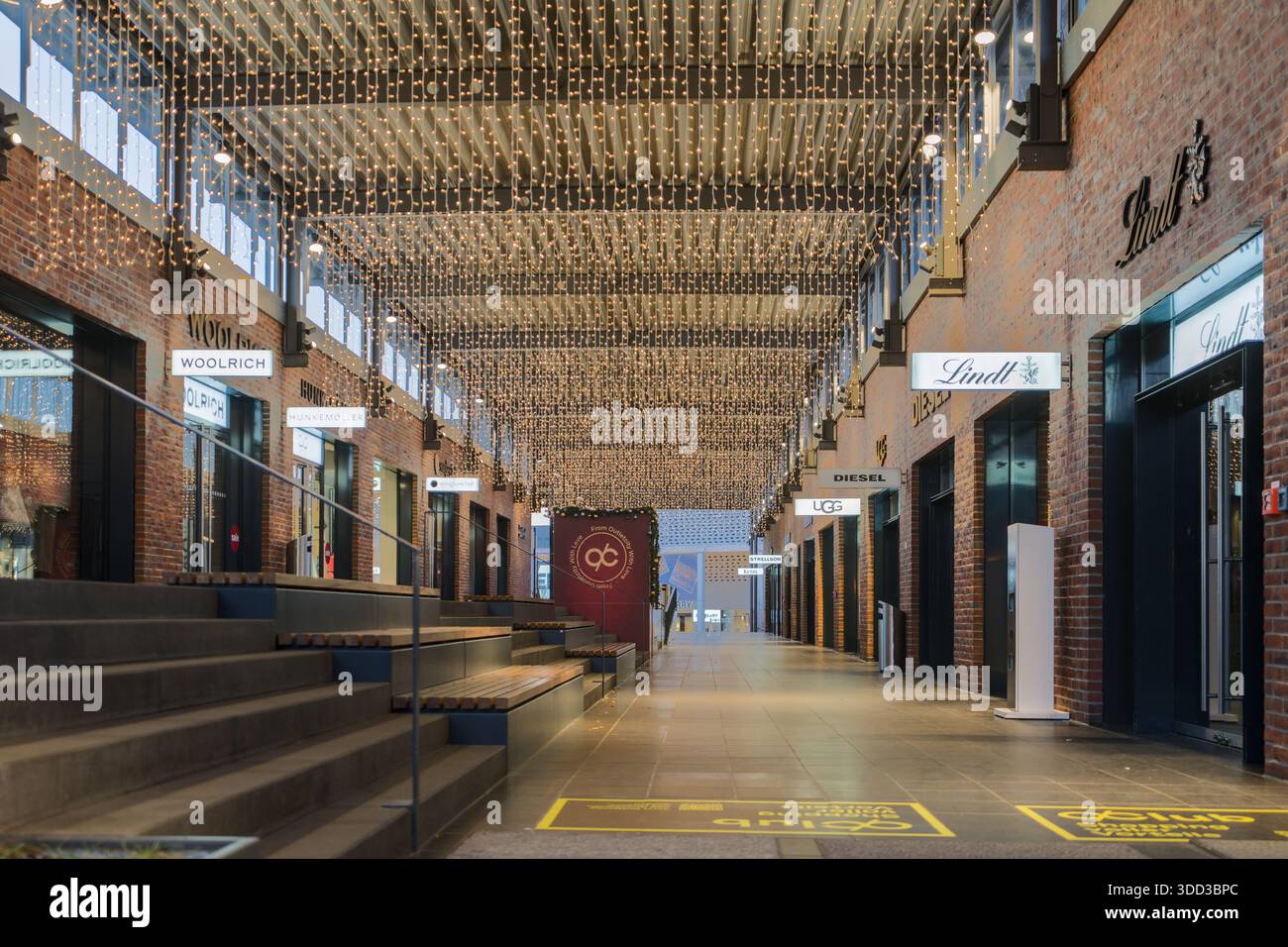 Festlich beleuchtete Einkaufspassage mit Backsteinmauern und verschiedenen Geschäften in einer lebendigen Outlet-Umgebung. Outletcity Metzingen Stockfoto