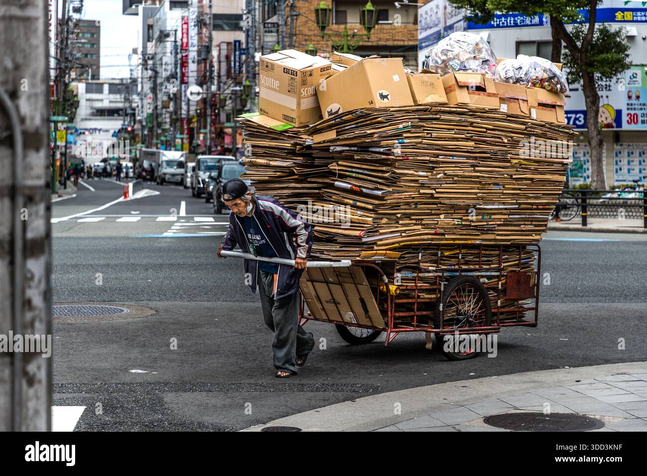 Ein Mann zieht einen stark beladenen Wagen, der mit Karton und Müllsäcken beladen ist, durch eine Straße in Osaka, Japan. Stockfoto