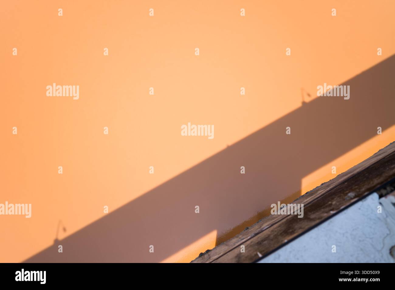 Der diagonale Schatten fällt über eine glatte pfirsichfarbene Wand, die von einem verwitterten Holzrand und einer strukturierten weißen Oberfläche durchzogen wird, wodurch eine minimalistische abstrakte Komposition in natürlichem Licht entsteht. Stockfoto