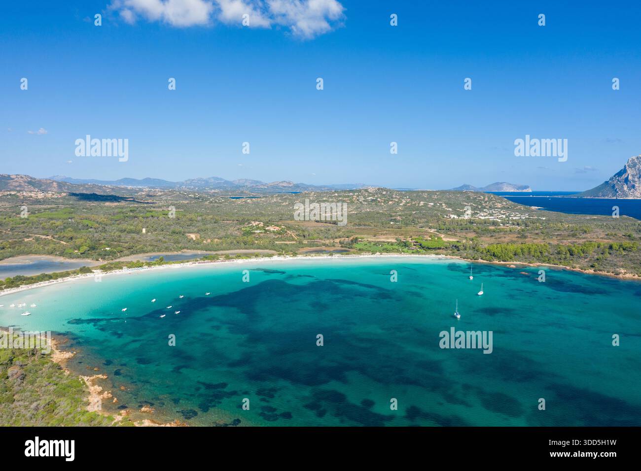 Blick aus einem Hochwinkel auf den Strand Cala Brandinchi auf Sardinien mit einem weiten Bogen aus türkisfarbenem Wasser, ankerbaren Booten und einer üppigen grünen Küste unter einem klaren Himmel mit weit entfernten Bergen. Stockfoto