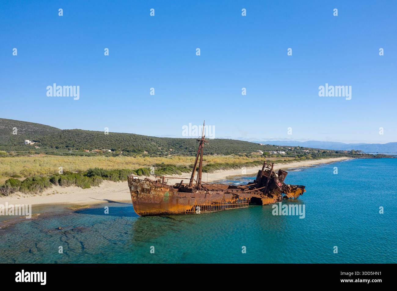 Ein verrostetes Schiffswrack liegt teilweise in klarem türkisfarbenem Wasser an einem Sandstrand in der Nähe von Gythio, umgeben von wilder Vegetation und fernen Hügeln unter einem leuchtend blauen Himmel. Stockfoto
