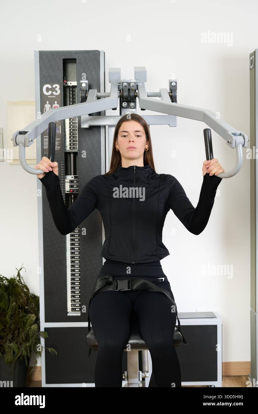 Junge Frau, die sich auf ihre Fitness-Routine konzentriert, auf einem Kraftgerät sitzt und in einem Fitnessstudio die Oberkörperkraft aufbaut Stockfoto