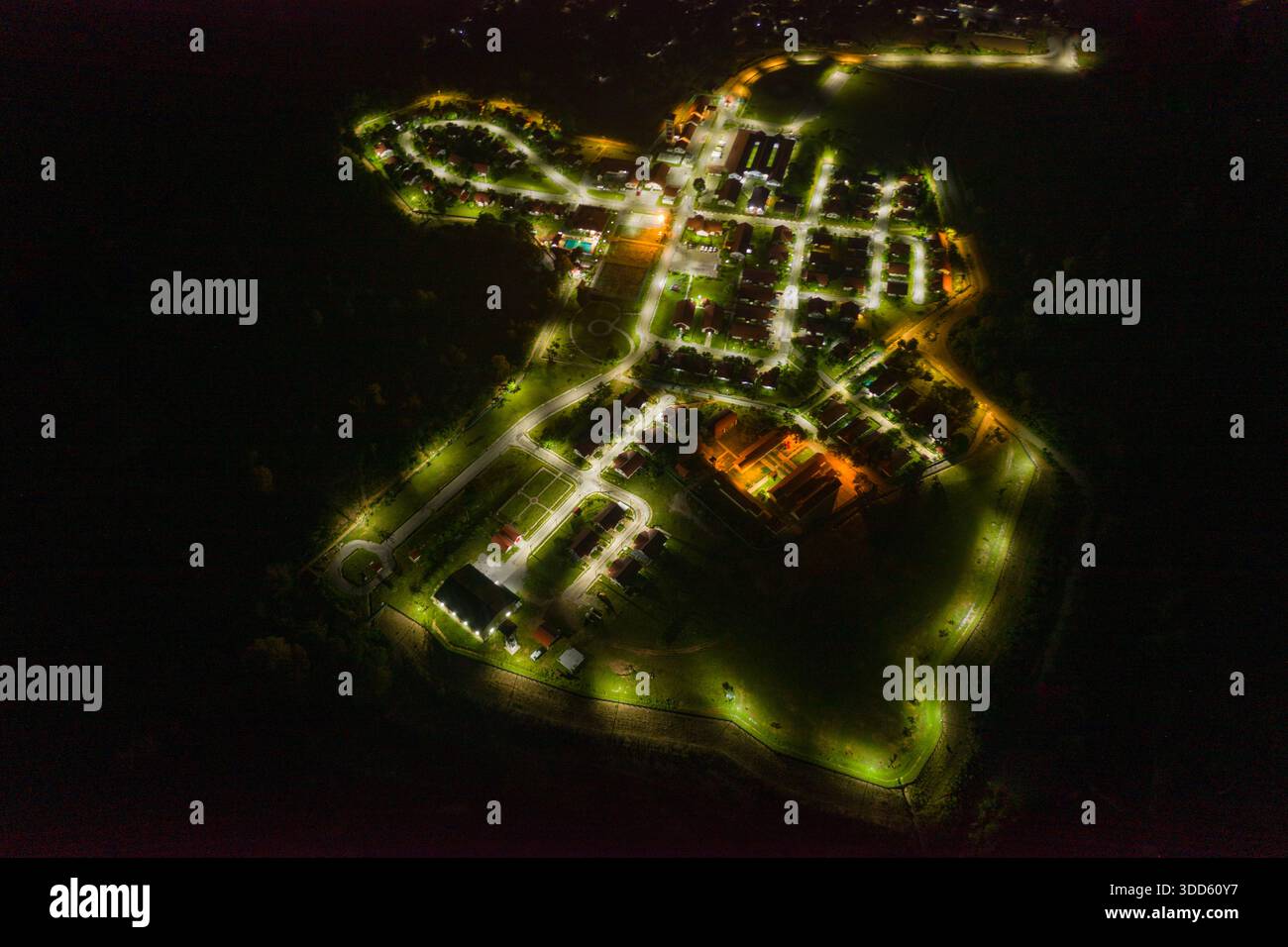 Drohnenperspektive, die ein kleines Dorf bei Nacht mit gewundenen Straßen und Wohngebäuden einfangen, die hell von Straßenlaternen beleuchtet werden. Die Szene zeigt geometrische Muster, kontrastierende Lichtquellen und das organisierte Layout der Siedlung, umgeben von Dunkelheit. Stockfoto