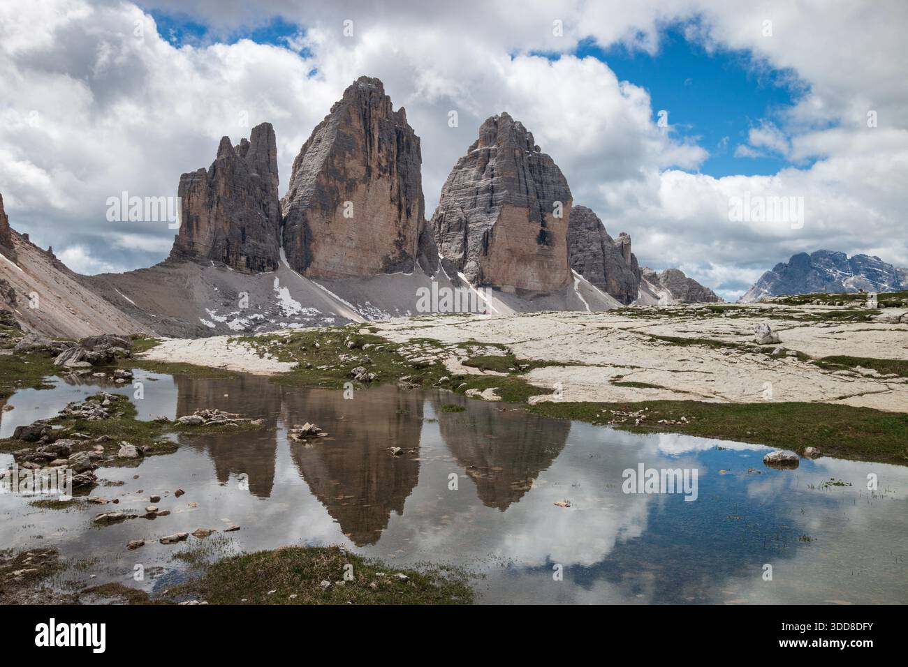 Niedriger Blickwinkel auf Tre Cime di Lavaredo in einer Pfütze; Cima Grande, Cima Ovest und Cima Piccola über hellem Kalksteingeröll vor blauem Himmel mit Wolken Stockfoto