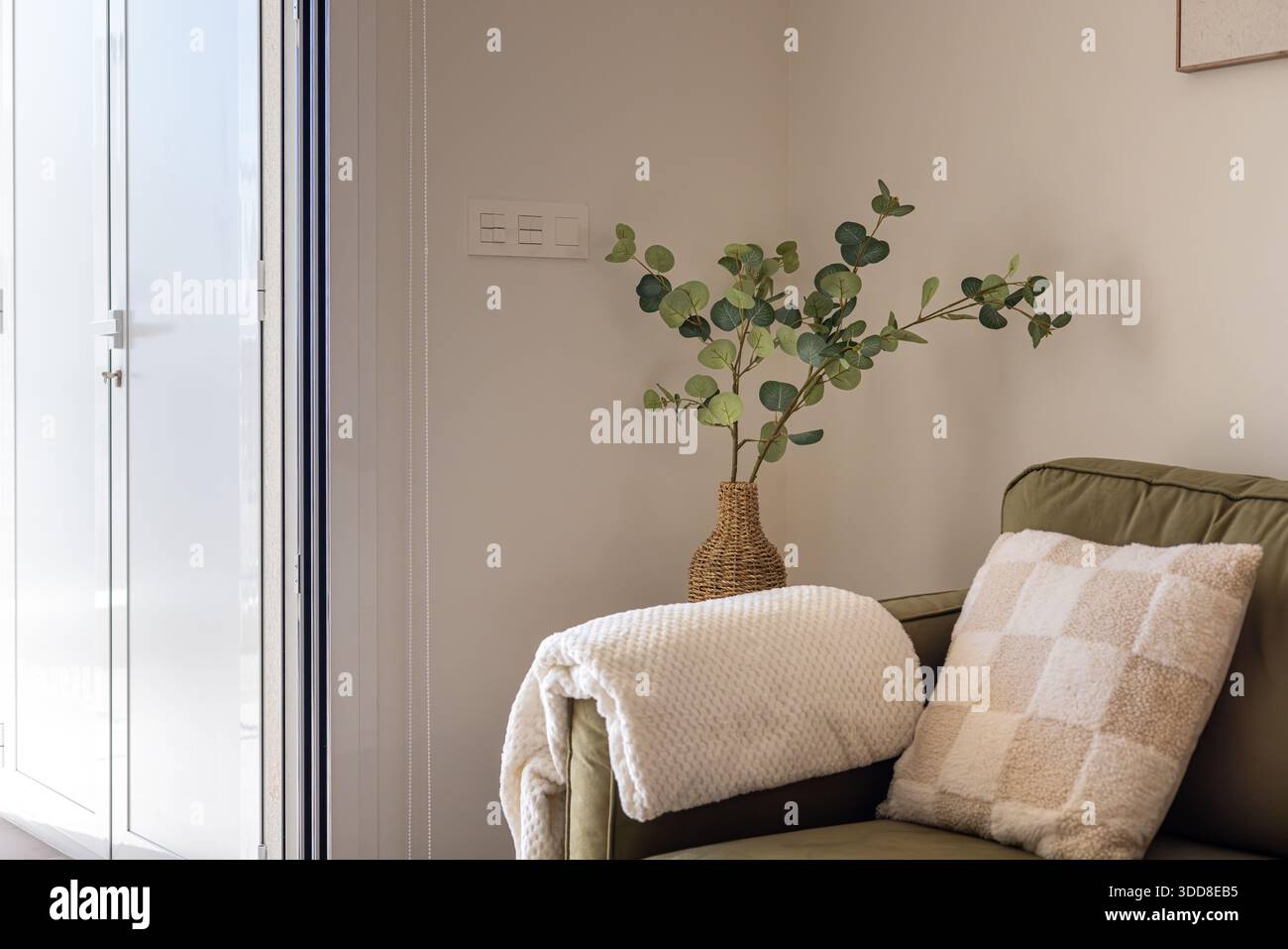 Sanft beleuchtete Ecke mit olivgrünem Sofa, Überwurfdecke, Kissen und Korbvase Stockfoto