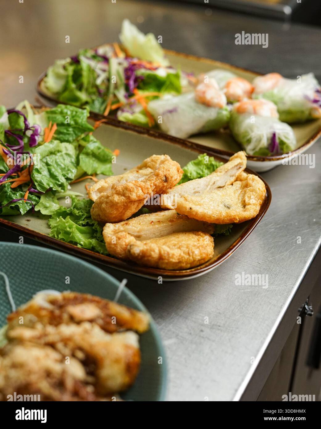 Verschiedene Gerichte, darunter Frühlingsbrötchen, Salat und gebratenes Hühnchen auf Metall, bieten vielfältige kulinarische Optionen. Stockfoto