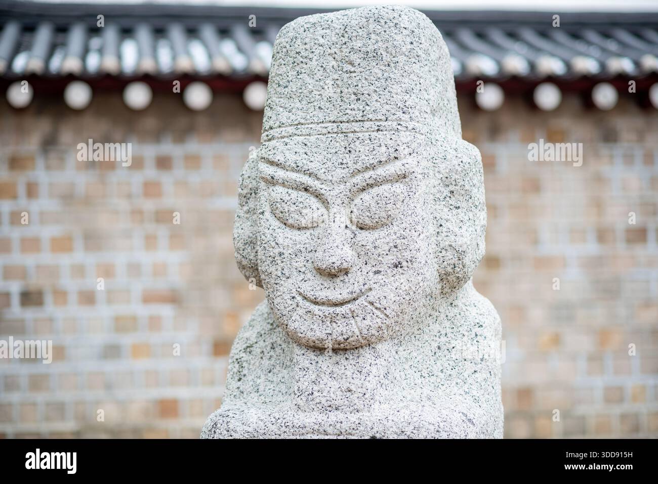 Eine Nahaufnahme einer koreanischen Steinstatue mit einem lächelnden Gesicht, mit traditionellem Kopfschmuck, vor einem verschwommenen Steinmauergrund Stockfoto
