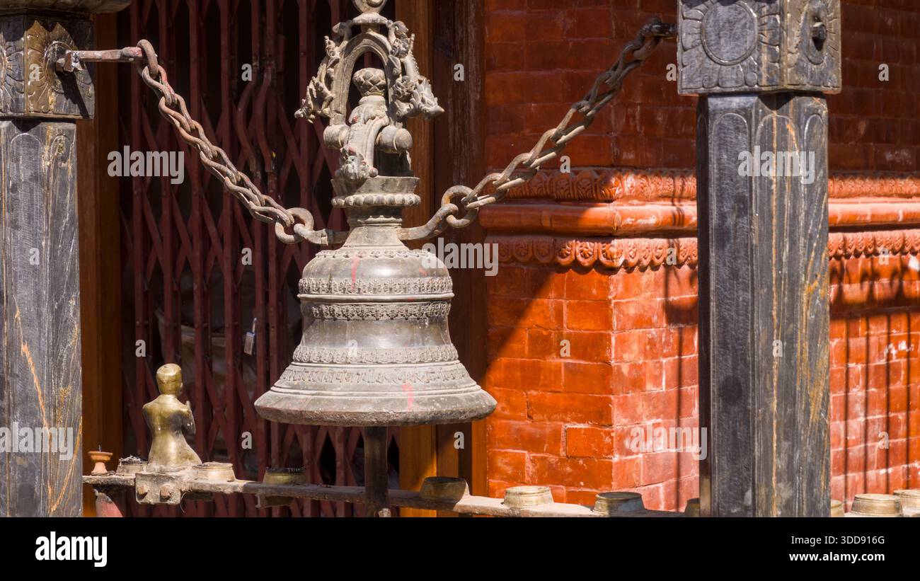 Aufwändig geschnitzte Metallglocke, die an Ketten an einer roten Ziegelwand in traditioneller Umgebung hängt. Stockfoto