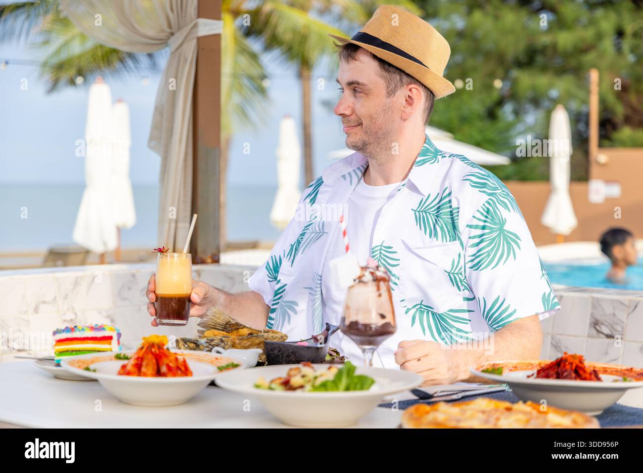 Mann genießt tropische Mahlzeiten im Strandrestaurant, trägt Strohhut und Palmhemd, umgeben von bunten Gerichten, Desserts und Getränken, unter sonniger Sonne Stockfoto