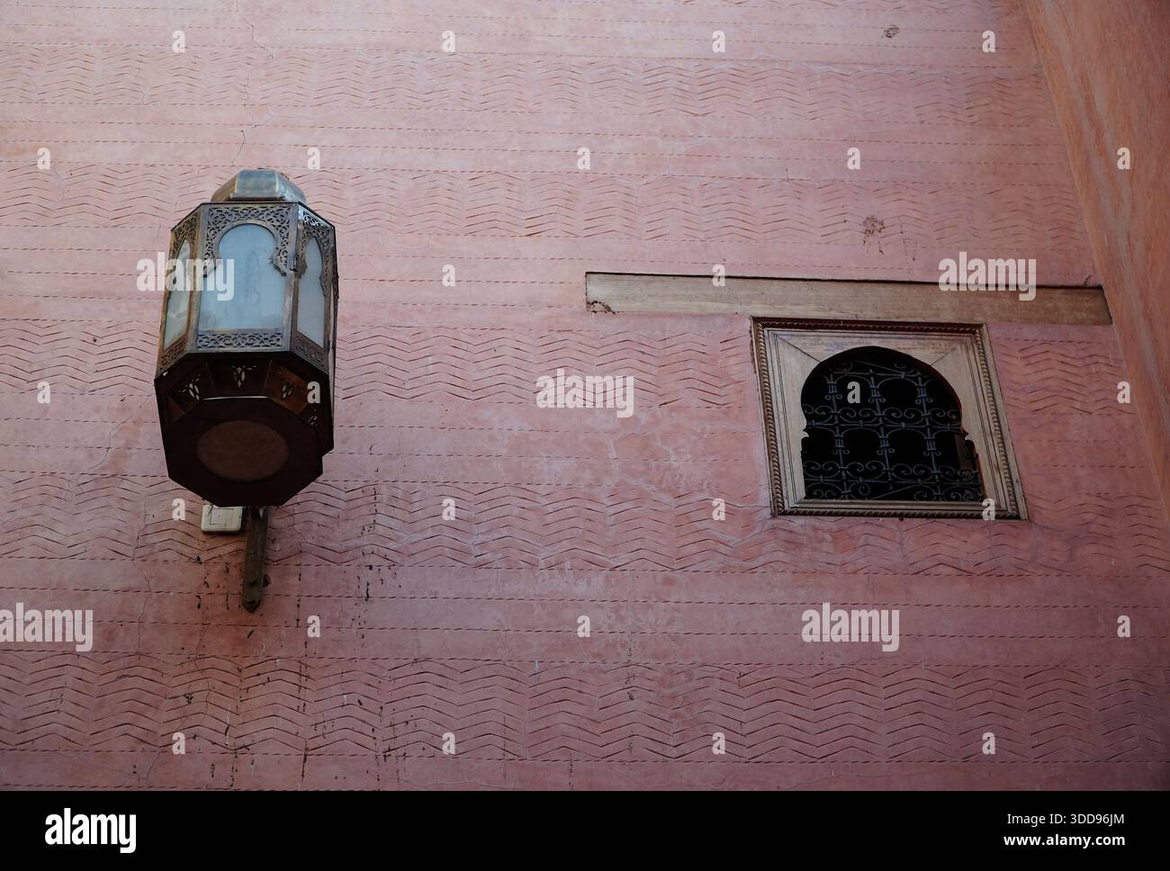 Strukturierte pinkfarbene Wand mit orientalischer Wandlaterne und kleinem Bogenfenster, geschützt durch einen schmiedeeisernen Grill, traditionelles architektonisches Ambiente. Stockfoto