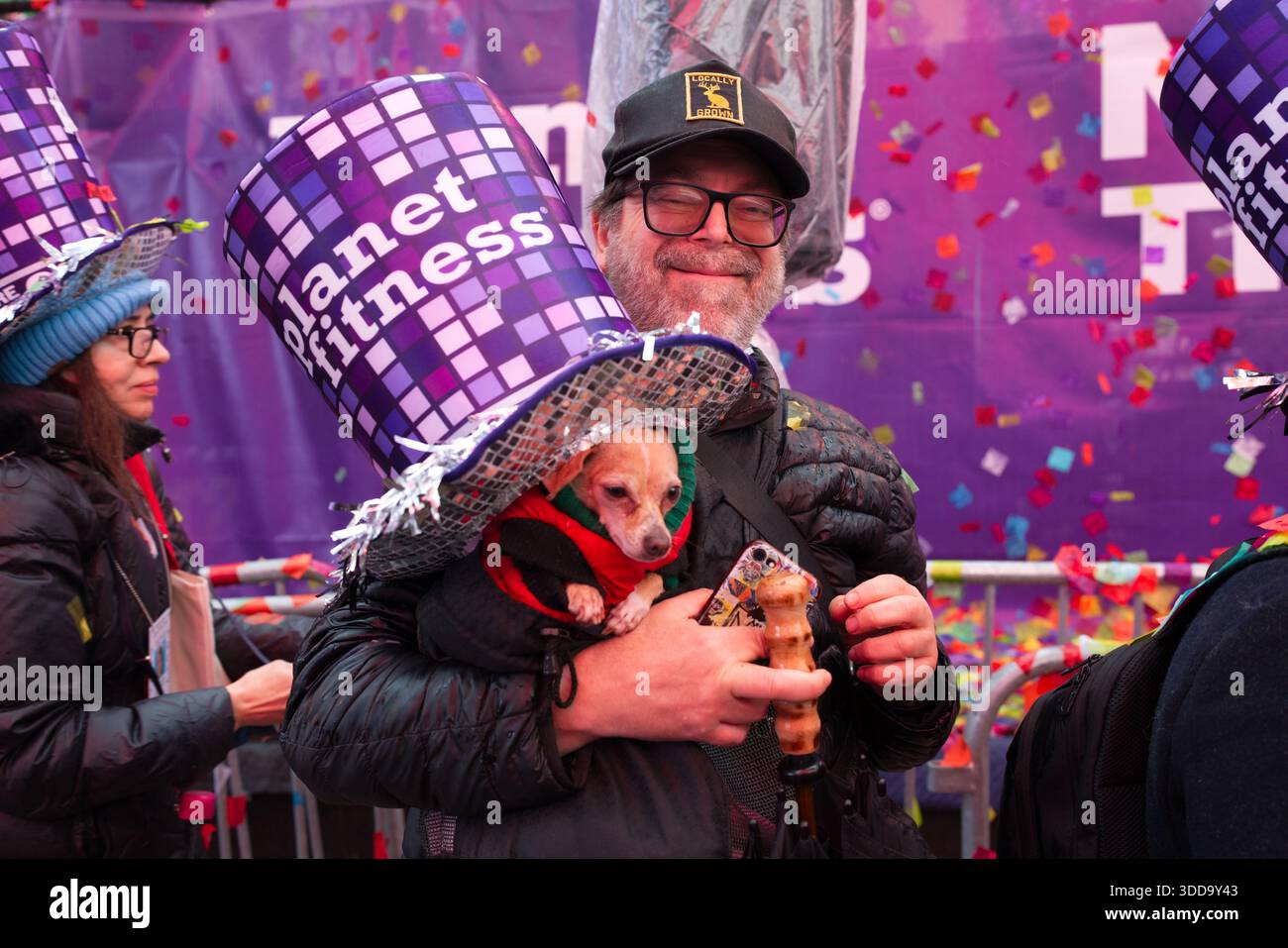 Ein Mann trägt einen großen, lila, glitzernden Hut mit dem Aufdruck „Planet Fitness“ und hält einen kleinen Hund. Konfetti fällt um sie herum. Stockfoto