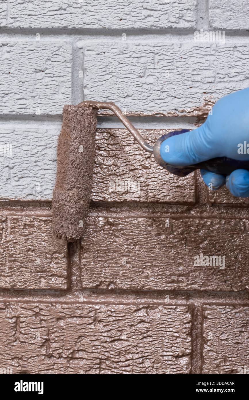 Person, die mit einer Malerwalze über eine graue Ziegelwand mit roségoldfarbener Metallic-Farbe malt. Machen Sie es sich selbst nach Hause Verbesserungskonzept. Stockfoto