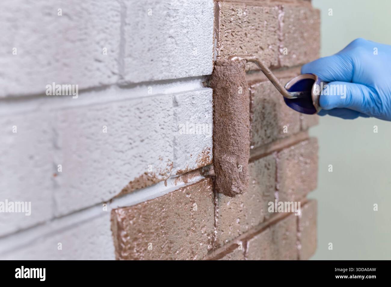 Person, die mit einer Malerwalze über eine graue Ziegelwand mit roségoldfarbener Metallic-Farbe malt. Machen Sie es sich selbst nach Hause Verbesserungskonzept. Stockfoto