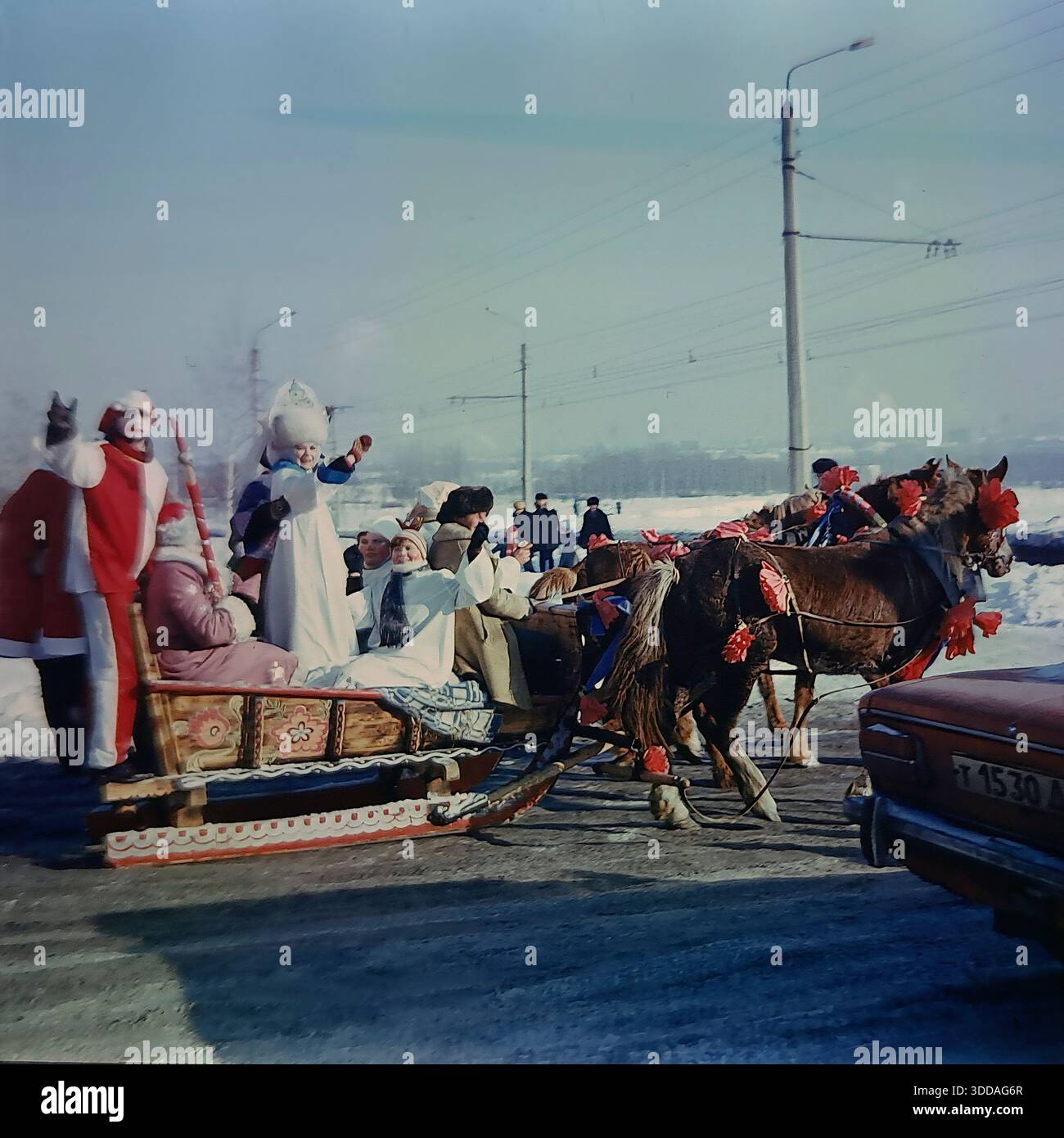 Slawjansk, Ukraine, UdSSR, 1980er Jahre Eine festliche Troika von Pferden zieht während der russischen Winterferien einen gemalten Holzschlitten durch das Artem-Mikroviertel. Schauspieler, die als Ded Moroz gekleidet sind, Snegurochka in einem Kokoshnik und helle Narren winken den Bewohnern zu. Der Folklorezug zieht vorbei an typischen sowjetischen Wohnhäusern und Fabrikschornsteinen. Ein Militärjeep der UAZ fährt in der Nähe. Die Szene steht in lebhaftem Kontrast zu traditionellen ländlichen Bräuchen und der modernen industriellen Stadtlandschaft der friedlichen Stadt. Stockfoto