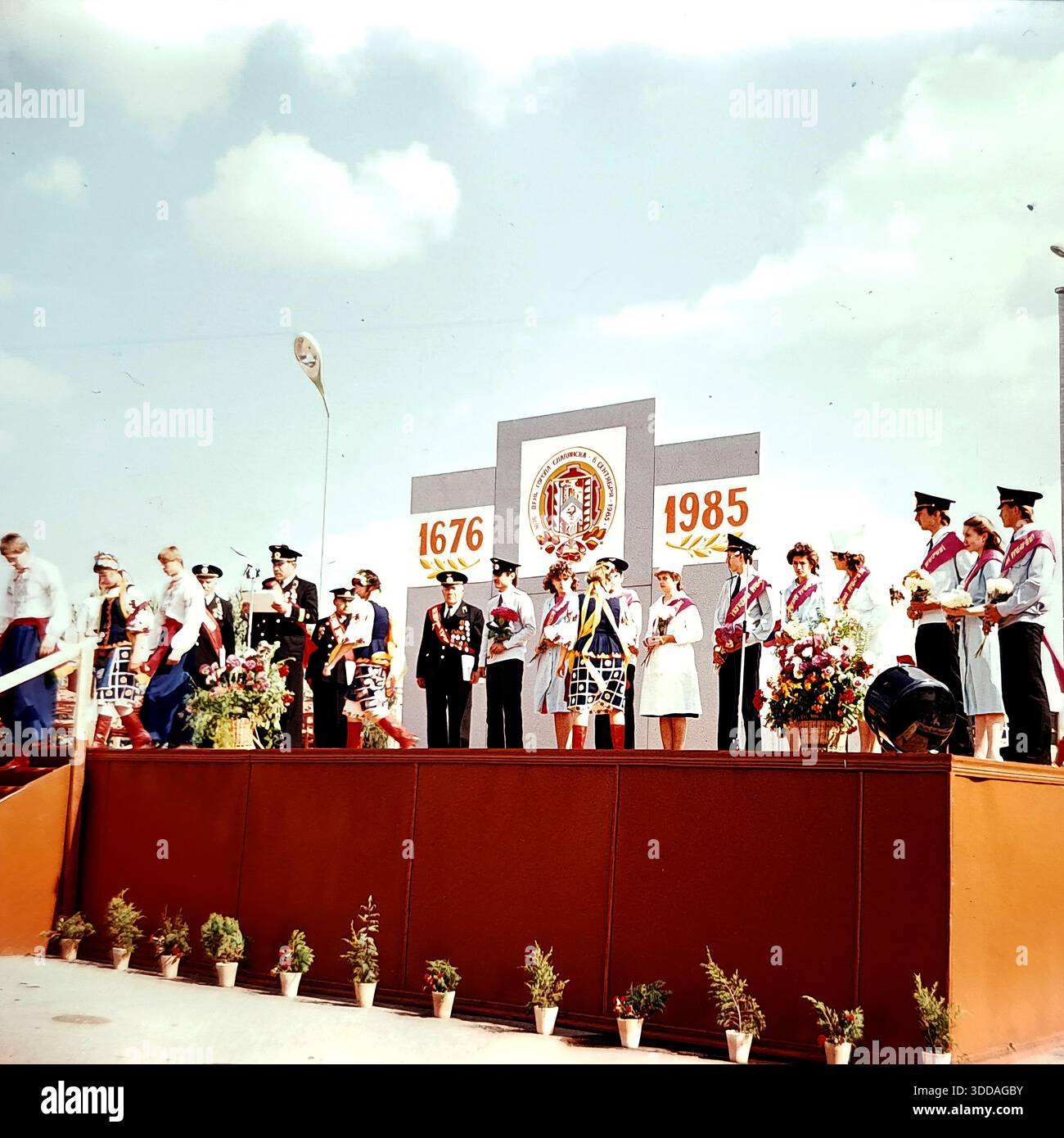 Slawjansk, Ukraine, UdSSR, 8. September 1985. Eine offizielle Zeremonie zum Stadttag findet auf einer Bühne am Oktyabrskoy Revolyutsii-Platz (heute Soborna-Platz) statt. Eine große Kulisse zeigt das Gründungsjahr 1676 und das laufende Jahr 1985. Veteranen des Zweiten Weltkriegs mit Medaillen und junge Ehrenbürger mit Schärpen stehen auf dem Podium, während ein Offizier am Mikrofon spricht. Tänzer in ukrainischen Trachten nehmen an der Veranstaltung teil. Dieses Archivbild fängt die patriotische Feier und Anerkennung der Bewohner der friedlichen Sowjetstadt ein. Stockfoto