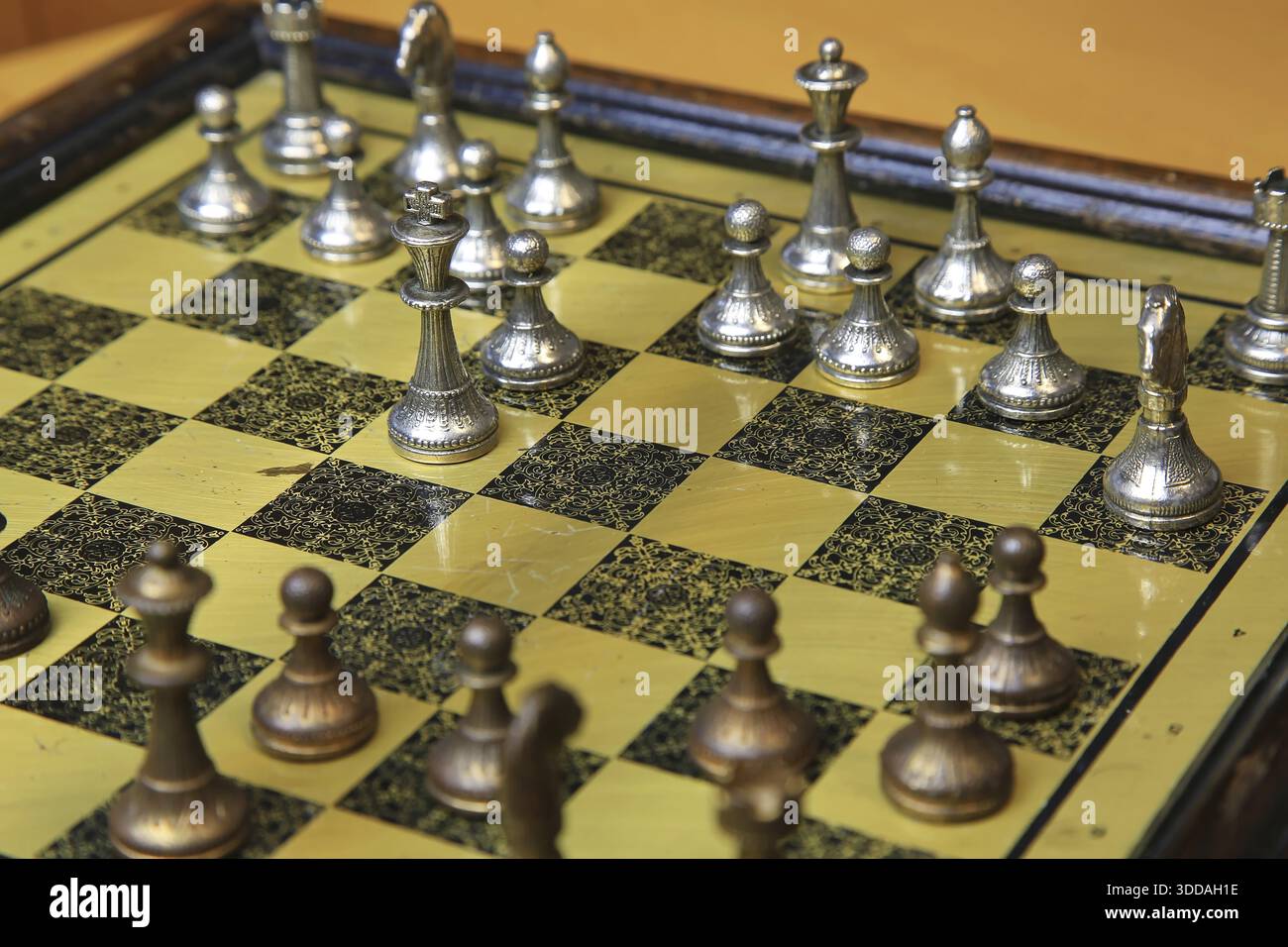 Ein feines Schachspiel auf einer gelben Pinnwand zieht die Aufmerksamkeit auf sich. Metallische Figuren in Silber und Bronze befinden sich in einer perfekten Ausgangsposition und reflektieren Stockfoto