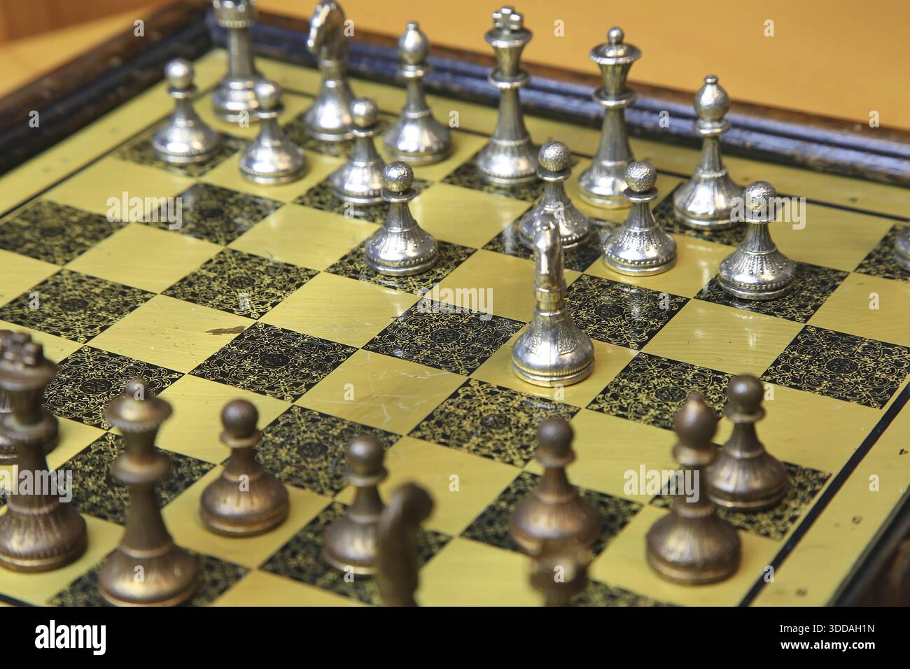Ein feines Schachspiel auf einer gelben Pinnwand zieht die Aufmerksamkeit auf sich. Metallische Figuren in Silber und Bronze befinden sich in einer perfekten Ausgangsposition und reflektieren Stockfoto