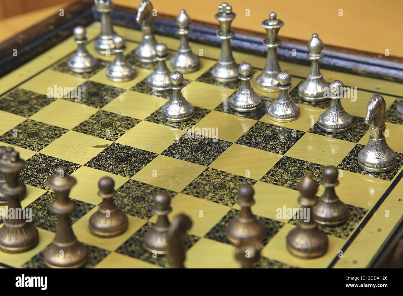 Ein feines Schachspiel auf einer gelben Pinnwand zieht die Aufmerksamkeit auf sich. Metallische Figuren in Silber und Bronze befinden sich in einer perfekten Ausgangsposition und reflektieren Stockfoto